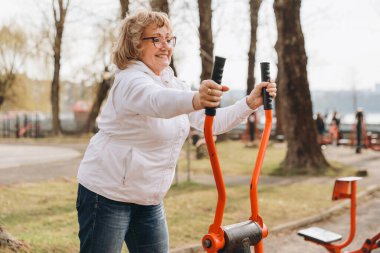 Elderly woman working out on elliptical trainer in a public park, enjoying healthy lifestyle and fitness