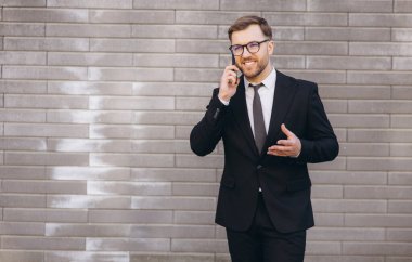 Smiling businessman having phone call wearing suit and tie standing near a gray brick wall