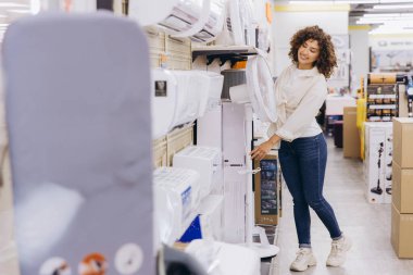 Young woman selecting a standing fan among various options in a home appliance store, considering comfort and cooling needs for summer