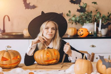 Young woman wearing a witch hat, happily carving pumpkins for a Halloween party in her cozy kitchen, embracing the festive spirit of autumn