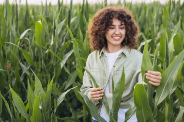 Smiling agronomist woman examining corn leaves and assessing crop health in a vibrant, cultivated corn field under the warm summer sun