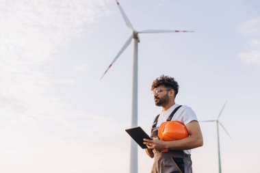 Engineer holding a tablet and helmet in a wind farm, focusing on sustainable energy solutions. Wind turbines in the background under a clear sky