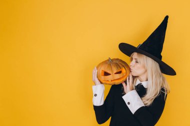 Halloween witch playfully blowing a kiss while holding a carved pumpkin, set against a vibrant yellow background, capturing festive spirit
