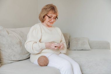 Elderly woman with glasses knitting on a comfortable sofa, enjoying her free time and retirement