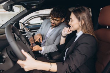 Car salesman showing new car features on tablet to businesswoman sitting in driver's seat at the car dealership