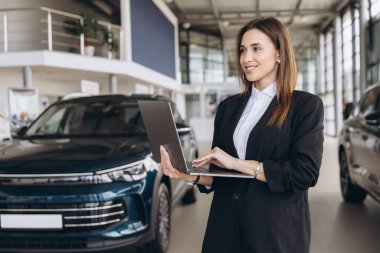 Smiling saleswoman using laptop, standing next to new car, inside modern car dealership showroom