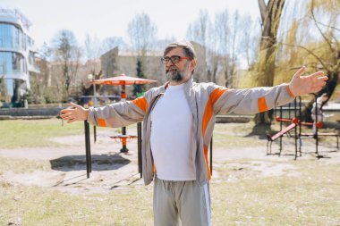 Bearded senior man wearing sportswear stretching arms in a calisthenics park, enjoying healthy lifestyle and wellness