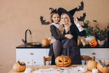 Mother and daughter smiling and wearing witch costumes while carving pumpkins and preparing for Halloween party in kitchen