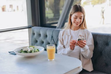 Happy customer using app on smartphone while eating healthy salad and drinking orange juice in modern restaurant