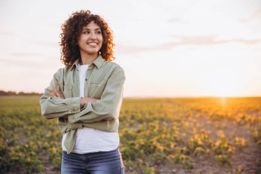 Confident female agronomist standing with crossed arms in a soybean field at sunset, smiling and looking away