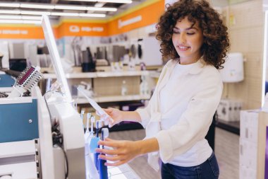 Smiling curly haired woman comparing electric toothbrushes in electronics store, choosing best toothbrush for her needs