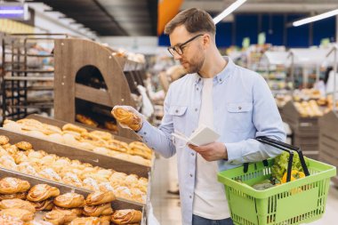 Customer selecting bakery products while holding shopping list and basket in supermarket