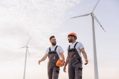 Two engineers walking and discussing near wind turbines, wearing uniforms and hardhats, in a sustainable energy context