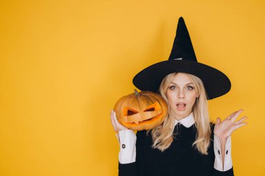 Young woman wearing witch costume holding carved pumpkin for halloween, surprised expression on yellow background