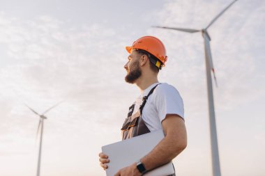 Engineer with an orange helmet and overalls is holding a laptop while inspecting wind turbines in a wind farm