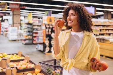 Young woman enjoying the aroma of a fresh apple while selecting groceries in a vibrant supermarket, embracing a healthy lifestyle