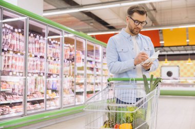 Man consulting his grocery list while pushing a shopping cart full of fresh produce in a supermarket