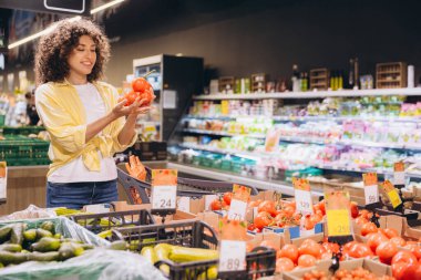 Smiling woman selecting tomatoes while grocery shopping in a brightly lit supermarket produce section, surrounded by fresh vegetables