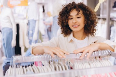 Curly haired woman shopping and selecting stylish clothes in a vibrant fashion store, enjoying the variety and making choices with a smile