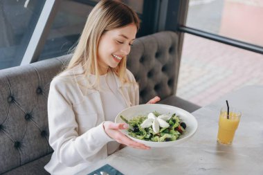 Young woman holding a plate of fresh salad with mozzarella cheese, smiling and enjoying her healthy meal in a restaurant