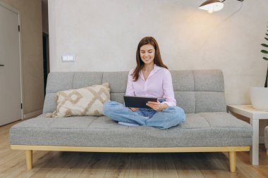 Smiling woman using digital tablet while sitting with crossed legs on comfortable sofa in cozy living room