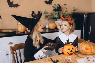 Mother wearing witch costume applying Halloween makeup on her daughter's face while preparing for the Halloween party
