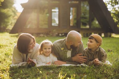 Happy family lying on blanket in the grass, enjoying a relaxing day in front of their modern a frame cabin
