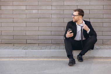 Exhausted businessman loosening his tie and using phone while sitting on the curb