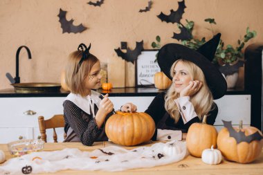 Mother wearing a witch hat and daughter joyfully carving pumpkins together for a festive Halloween celebration at home in the kitchen