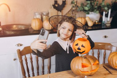 Girl taking a selfie with her smartphone and a carved pumpkin while wearing a witch costume for Halloween