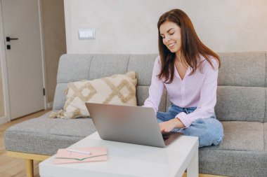 Happy businesswoman using laptop, typing on keyboard, enjoying remote work from home, sitting on comfortable sofa in living room