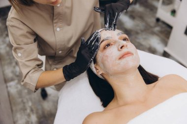Beautician wearing black gloves applying a facial mask on the face of a young woman lying on a bed in a beauty salon