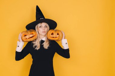 Young woman wearing witch costume holding two carved pumpkins on yellow background for halloween party