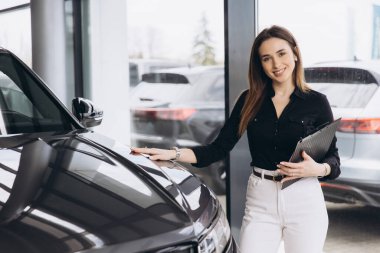 Saleswoman holding clipboard presenting new car to customer in car dealership