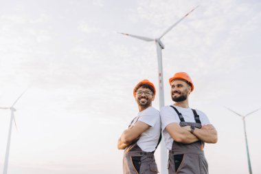 Two smiling male engineers with crossed arms standing back to back in a wind turbine power plant, embodying renewable energy and sustainability