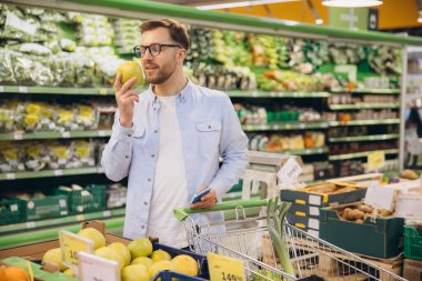 Customer smelling a green apple while holding a shopping list and pushing a shopping cart in a supermarket