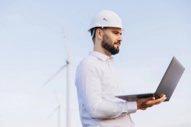 Engineer is holding a laptop and inspecting wind turbines in a field, contributing to sustainable energy development