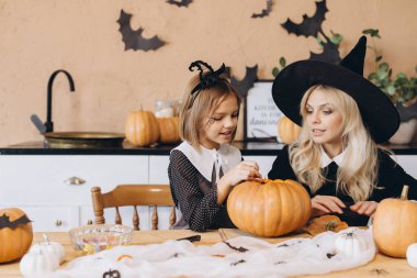 Mother wearing witch hat and daughter carving pumpkins for Halloween party, home kitchen decorated with bats and spiders