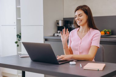 Young woman waving cheerfully at the camera while engaging in a video call on her laptop in a cozy home office setting