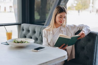 Young woman enjoying a healthy lunch of salad and orange juice while reading a book in a modern cafe, embracing a peaceful moment