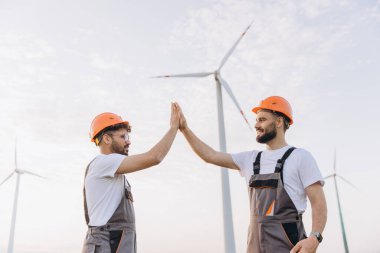 Two engineers in hard hats high fiving at a wind farm, symbolizing teamwork and success in sustainable energy production