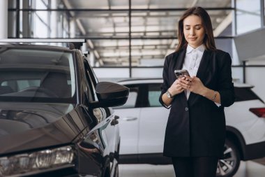 Saleswoman using a smartphone while standing beside a car in a dealership, engaging with customers and navigating the digital marketplace