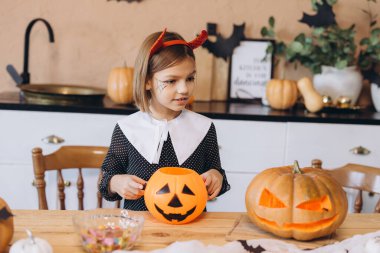 Girl with devil headband and spiderweb makeup holding a jack o' lantern bucket with candies, sitting at wooden table with carved pumpkins in decorated kitchen
