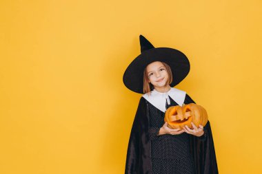 Little girl wearing a whimsical witch costume, joyfully holding a carved pumpkin against a bright yellow background, celebrating Halloween