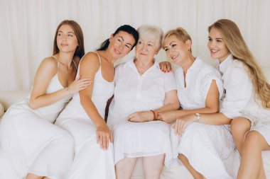 Four generations of women of the same family sitting together on a white sofa, wearing white clothes, looking away from camera