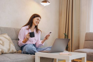 Smiling woman sitting comfortably on sofa, holding coffee mug and using smartphone while working on laptop at home
