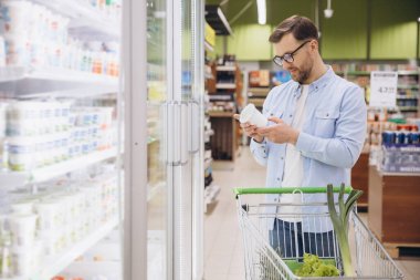 Man pushing shopping cart and reading label of yogurt in refrigerated section at supermarket