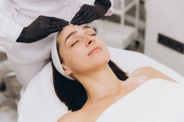 Beautician gently applying cream on a young woman's forehead during a facial treatment in a professional spa setting, promoting skincare and relaxation