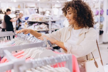 Smiling curly haired woman selecting clothes in a fashion store, enjoying shopping and choosing new outfits
