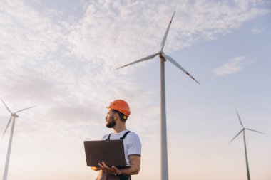 Engineer with safety helmet holding laptop inspecting wind turbines in a wind farm at sunset for clean energy